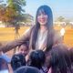 A young woman stands smiling at the camera in an outdoor space whilst a group of young children gather round her to hug her.