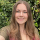 Amy has long brown hair with pink ends. She wears a green top and stands in front of a hedge.
