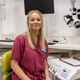 Lucy has blonde hair and wears red scrubs. She sits in a chair with dental phantom heads behind her.